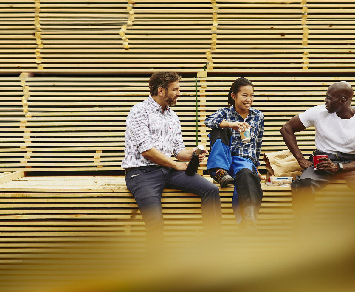 Three colleagues having a laugh during a lunch break while sitting on a pile of wood.
